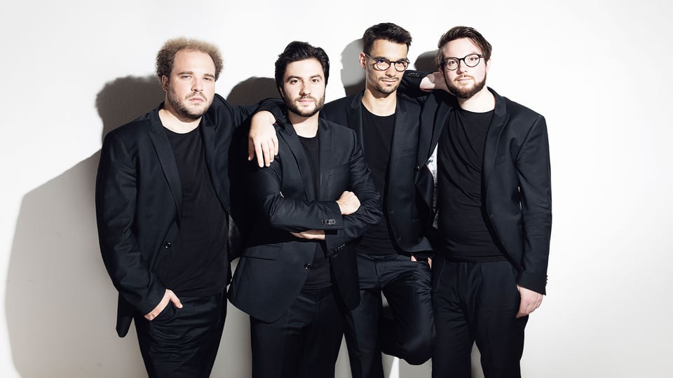 Goldmund Quartett mit vier Musiker:innen in schwarzen Anzügen vor hellem Hintergrund, nebeneinander stehend, teilweise mit verschränkten Armen oder aufgelegten Händen, ohne Instrumente im Bild sichtbar.