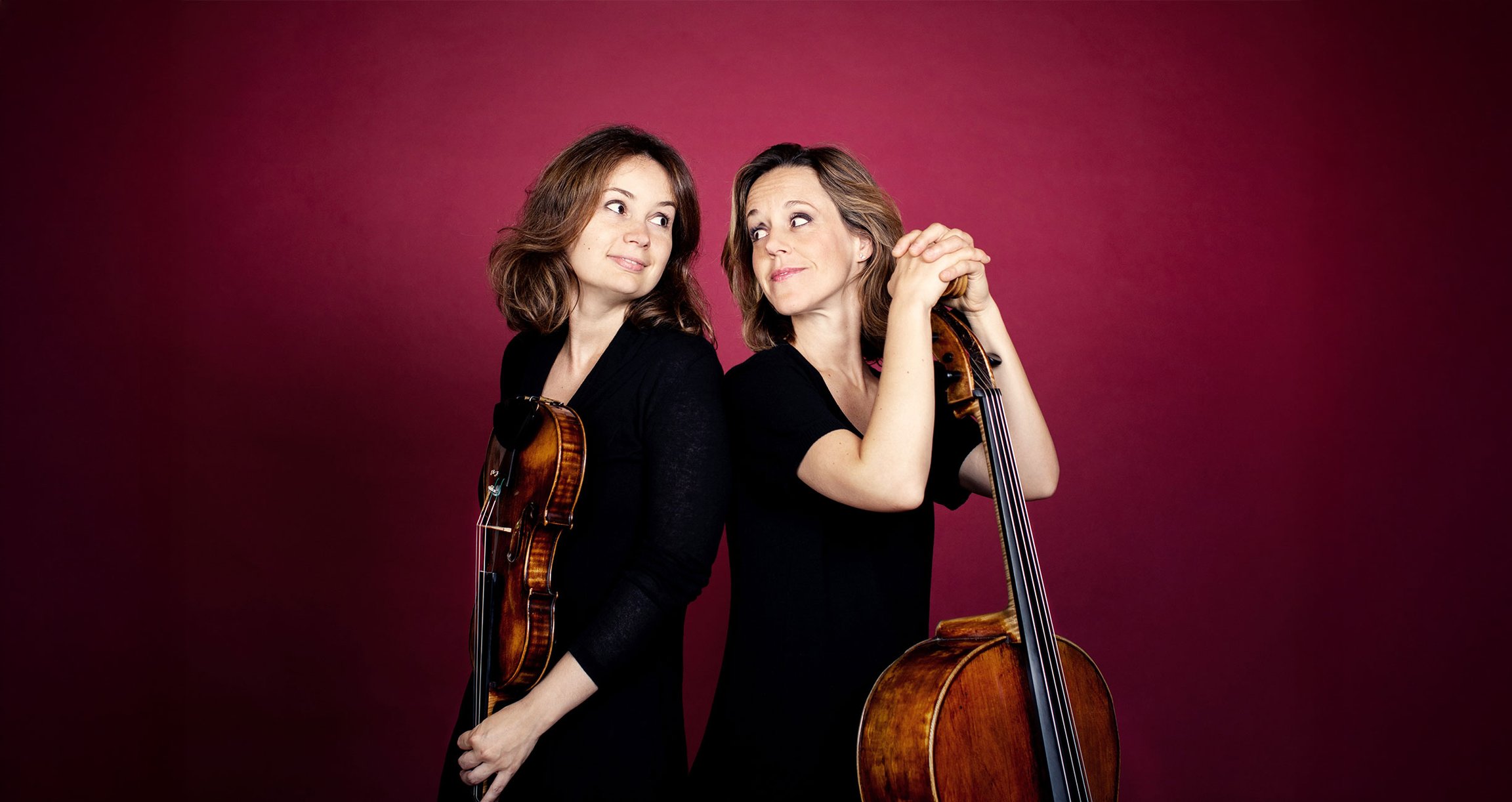 Sol Gabetta und Patricia Kopatchinskaja Rücken an Rücken vor einem roten Hintergrund mit ihren Instrumenten