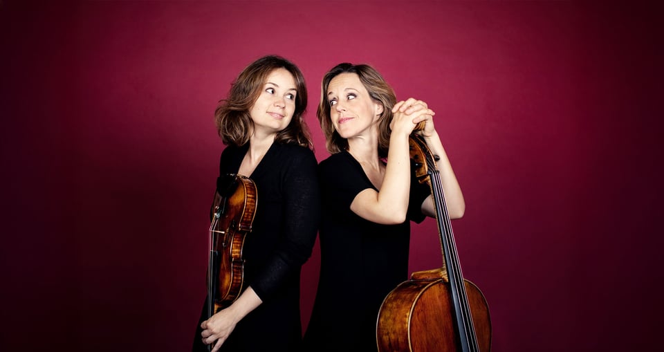 Sol Gabetta und Patricia Kopatchinskaja Rücken an Rücken vor einem roten Hintergrund mit ihren Instrumenten