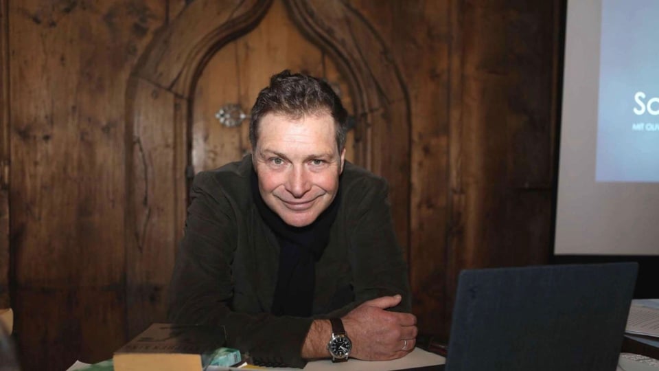 Oliver Pötsch in dunklem Sakko und schwarzem Rollkragenpullover, die auf einem Tisch vor einer geschnitzten Holzwand lehnt, neben Büchern und einem Laptop in einem holzvertäfelten Raum