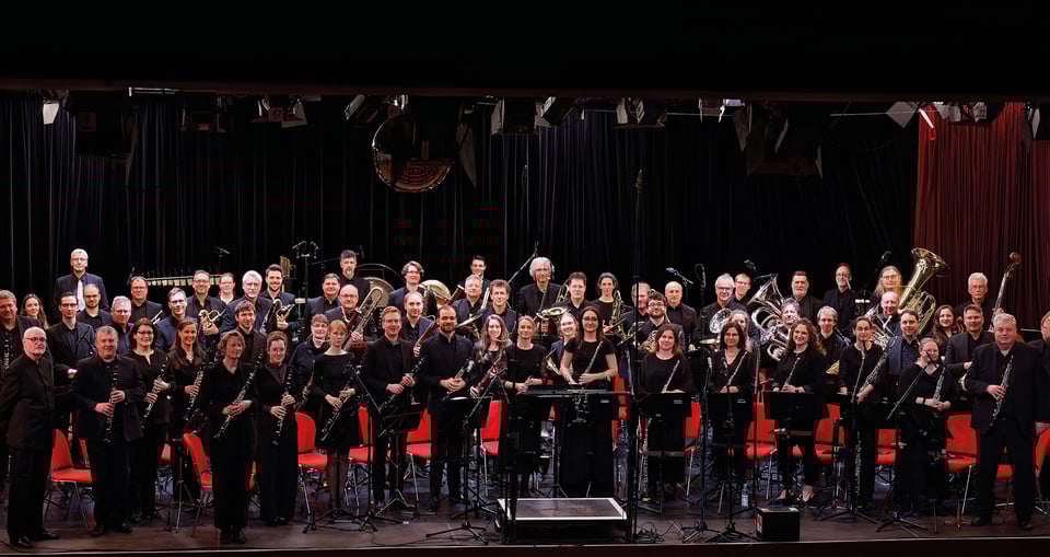 Foto eines Orchesters, alle Musiker in Schwarz gekleidet, hinter jedem steht ein roter Stuhl.