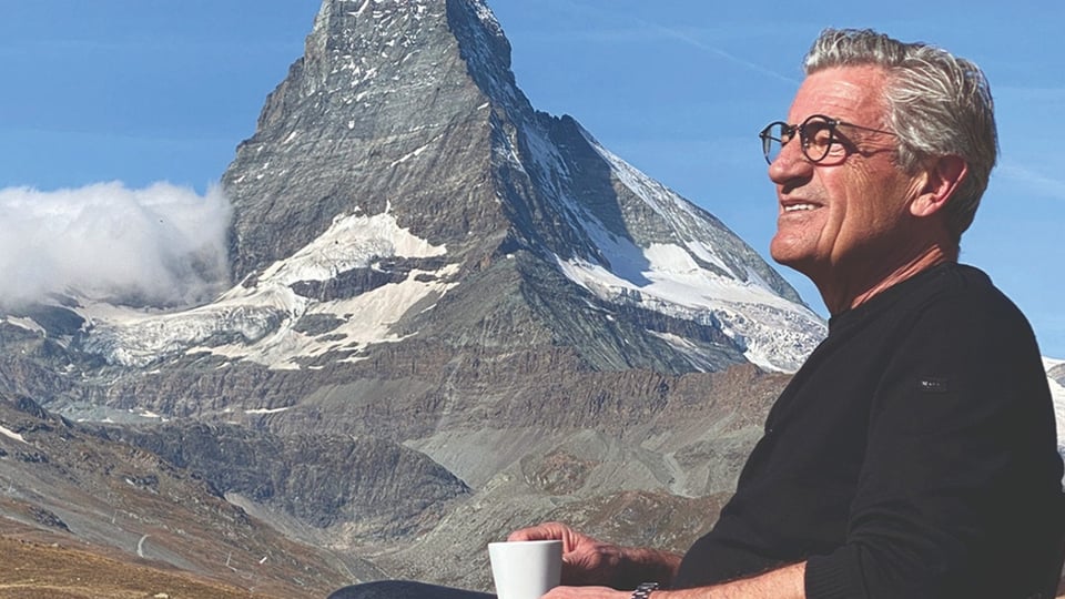Eine Person mit Brille in schwarzer Kleidung mit Kaffetasse in der Hand vor einer Bergmassiv