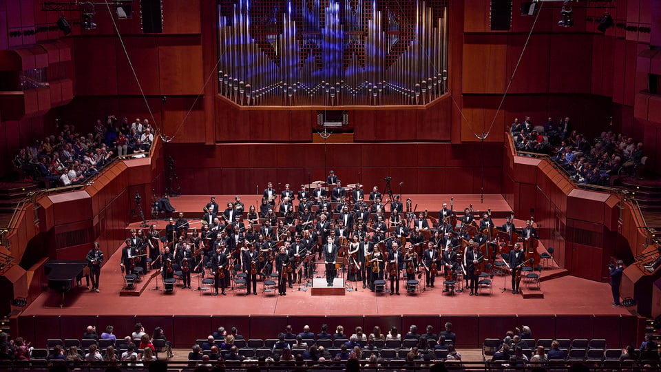 Gesamtaufnahme des Münchner Festspielorchesters auf der Bühne eines Konzertsaals mit Dirigentenpodest, zahlreichen Musikerinnen und Musikern sowie Publikum auf den Rängen 