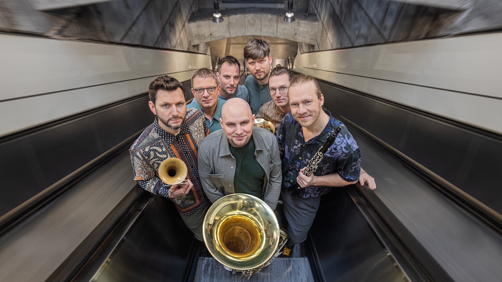 Sieben Musiker stehen mit Blas- und Holzblasinstrumenten auf einer Rolltreppe und blicken in die Kamera.