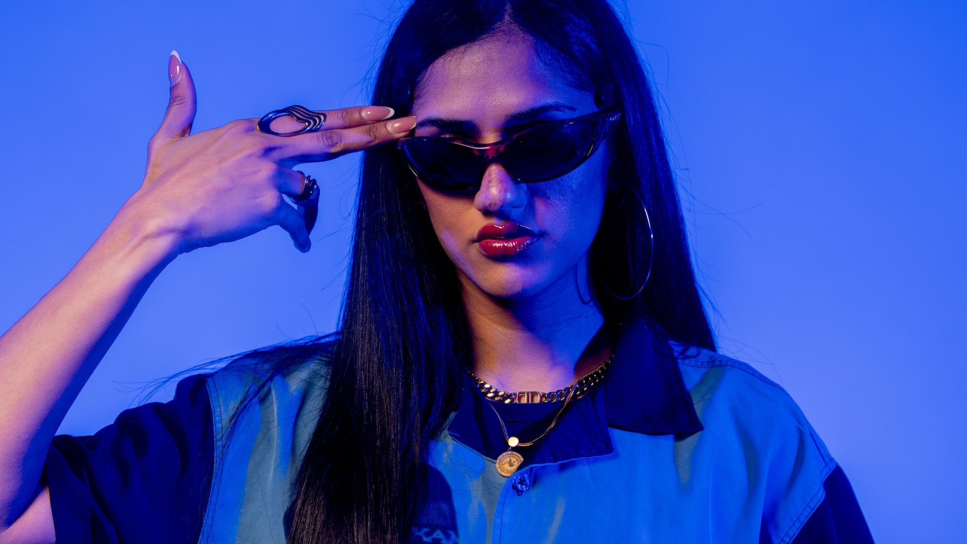 Porträtfoto von Wa22ermann mit langen dunklen Haaren, schmaler Sonnenbrille und blauem Hemd vor blau violett beleuchtetem Hintergrund, die Hand in Form einer Pistole an die Schläfe haltend.