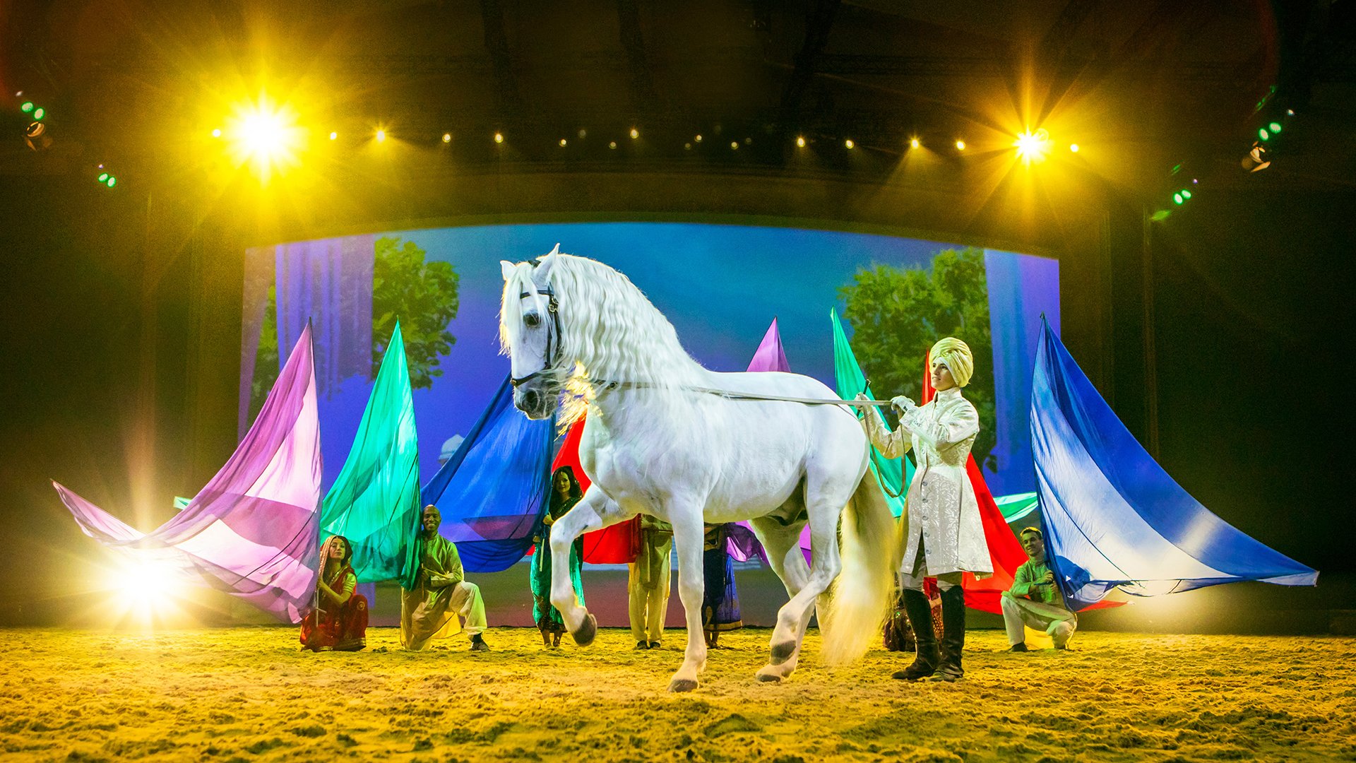 Weißes Pferd schreitet über eine beleuchtete Bühne, geführt von einer Person; im Hintergrund sitzen weitere Mitwirkende mit farbigen Stoffbahnen vor großer Bühnenprojektion und warmem Licht.