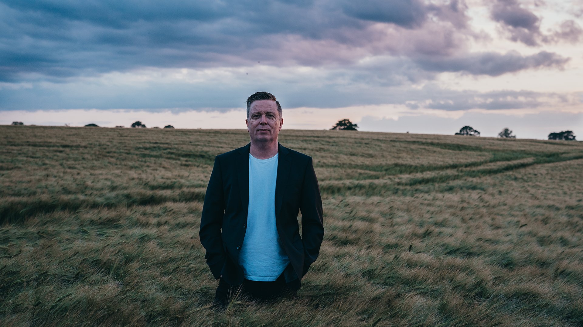 Eventmotiv von Dirk Maassen: Eine Person steht mittig in einem weiten, grasbewachsenen Feld unter einem dramatisch bewölkten Himmel. Die Person trägt dunkles Sakko über hellem Shirt, Hände in den Taschen. Natürliche Landschaft, gedämpftes Tageslicht; ruhiges, cineastisches Porträtmotiv zur Live‑Konzertankündigung von Dirk Maassen.