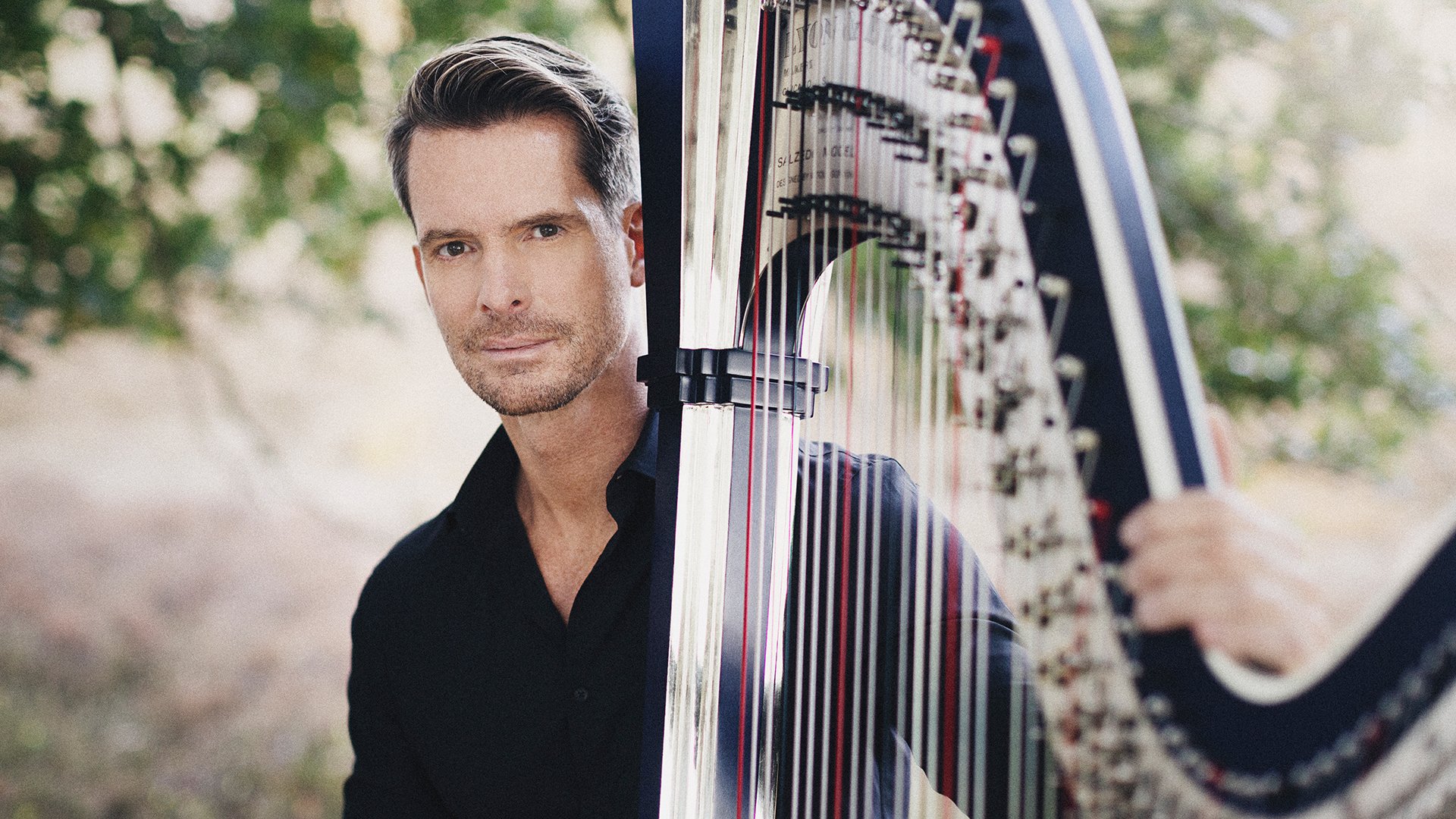 Promobild für das Event ‚Münchener Kammerorchester / Maistre‘: Eine Person in dunkler Kleidung steht im Freien neben einer großen Konzertharfe, deren Saiten und Rahmen im Vordergrund deutlich sichtbar sind. Im Hintergrund sind Bäume und unscharfe Natur zu erkennen