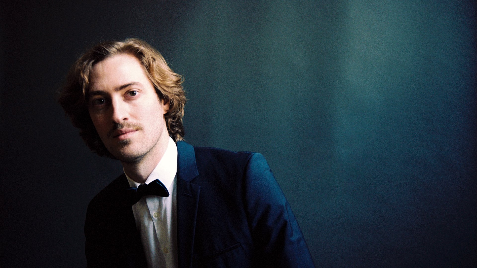 Der Pianist Alexander Maria Wagner in weißem Hemd dunkelblauem Sakko mit Fliege vor petrolfarbenem verschwommenen Hintergrund