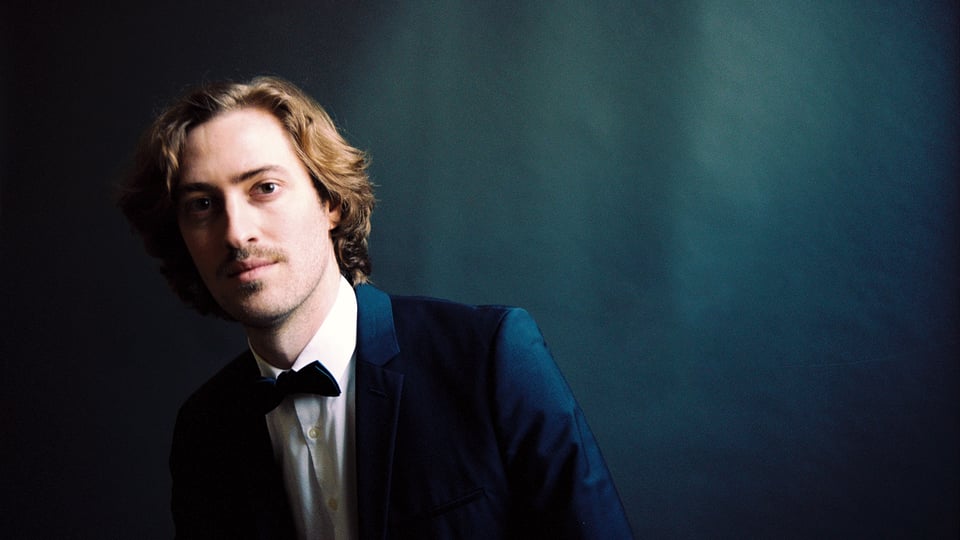 Der Pianist Alexander Maria Wagner in weißem Hemd dunkelblauem Sakko mit Fliege vor petrolfarbenem verschwommenen Hintergrund