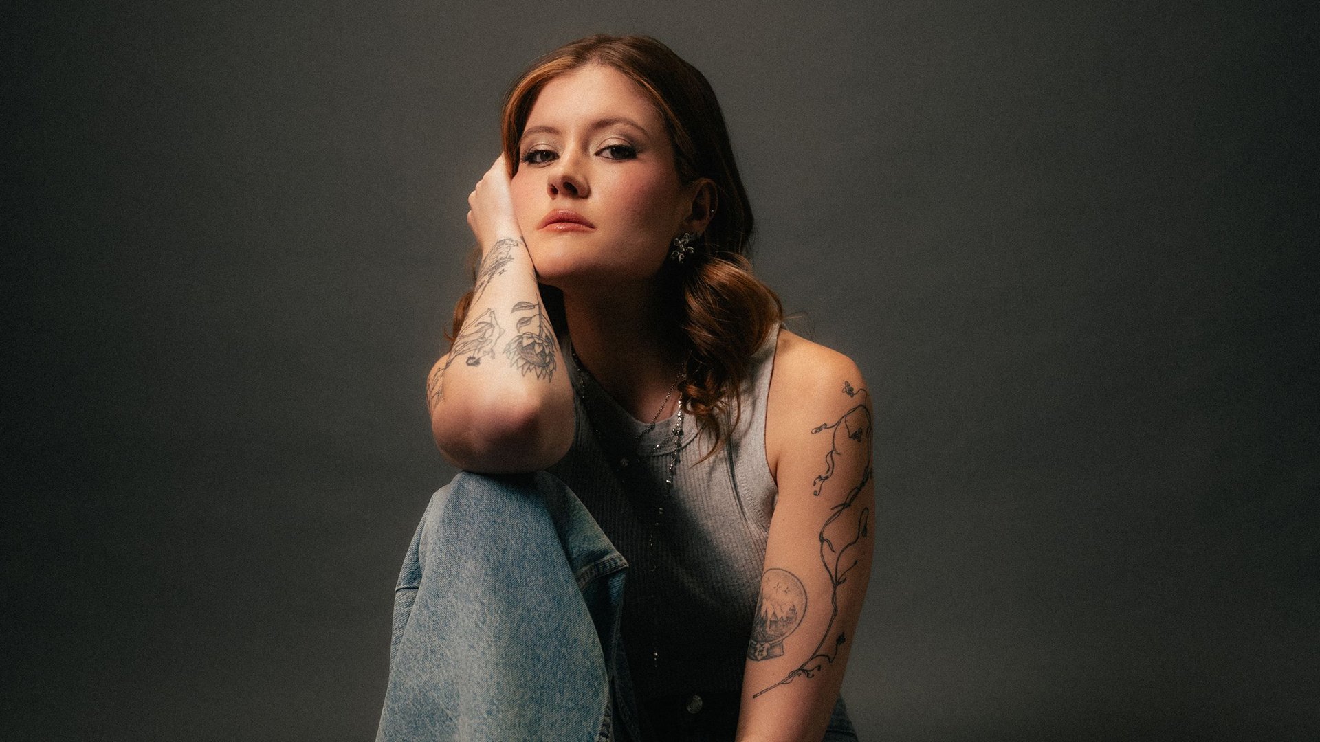 Madeline Juno in Jeans sitzt mit dem Ellbogen auf das Knie gestützt und blickt in die Kamera, vor grauen Hintergrund