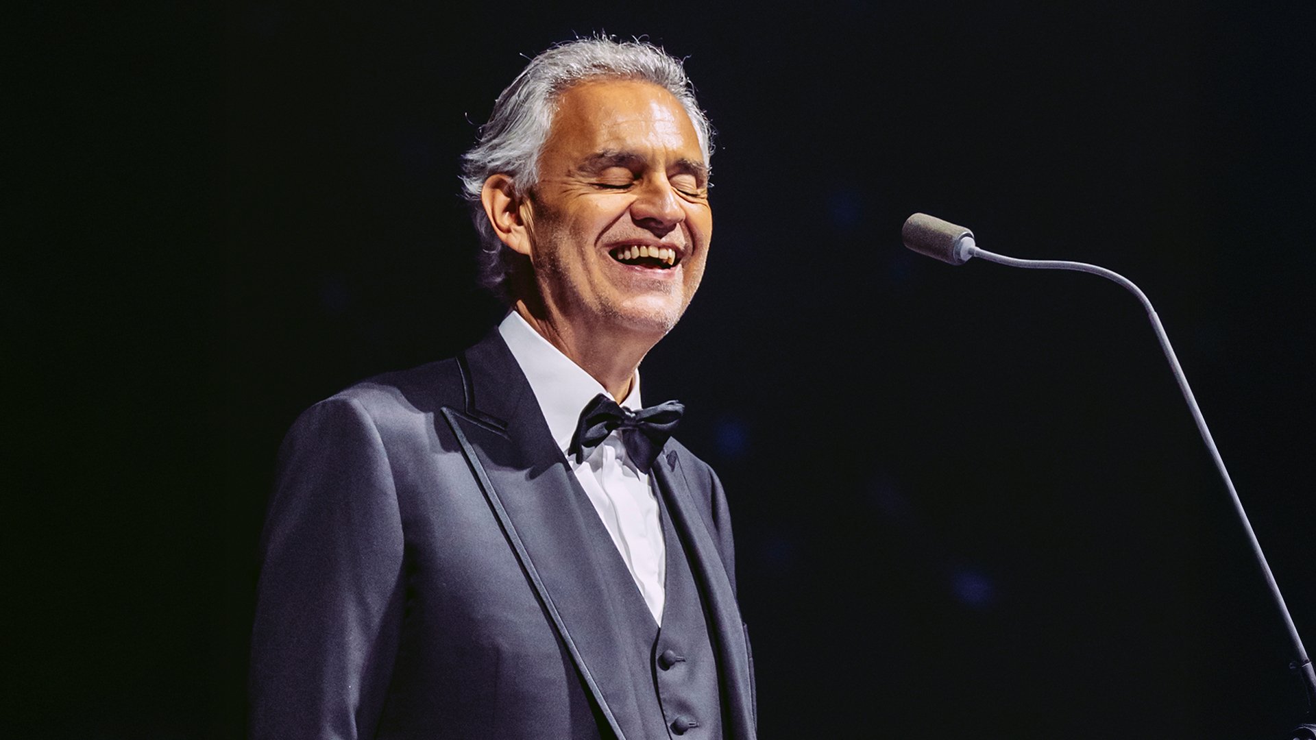 Der Künstler Andrea Bocelli in einem dunklen Anzug mit weißem hemd und Fliege, am Mikrofon stehend, vor schwarzem Hintergrund