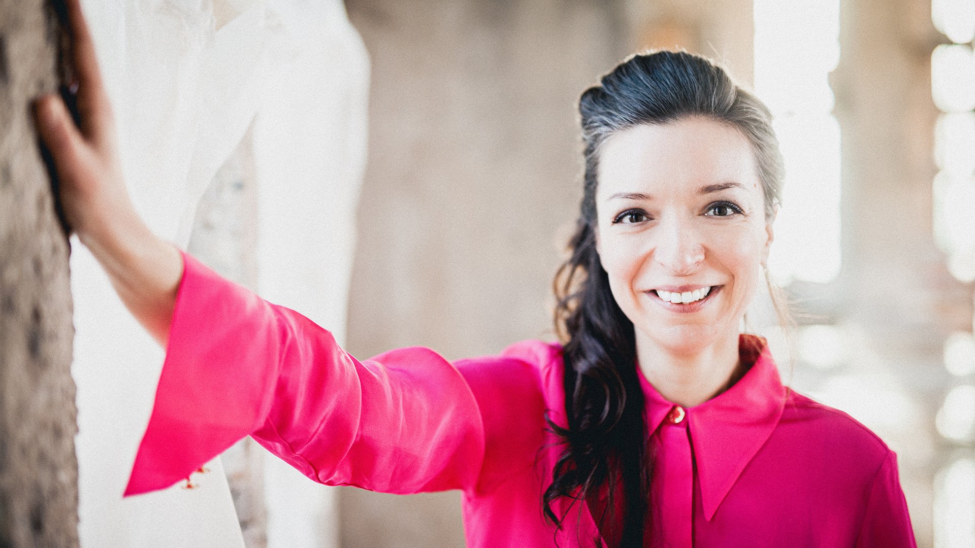 Yulianna Avdeeva in leuchtend pinker Kleidung, an eine Wand gelehnt, vor hellem Hintergrund.