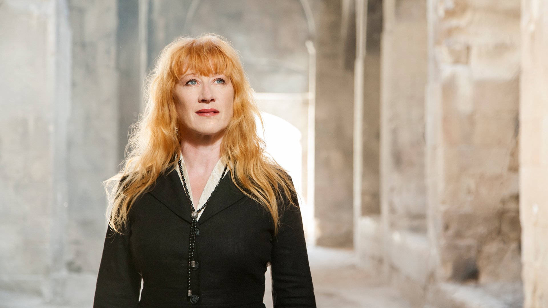 Loreena McKennitt in schwarzer Kleidung und offenen Haaren leicht nach oben blickend vor einer historischen Kulisse aus Mauern und Torbögen aus hellem Stein