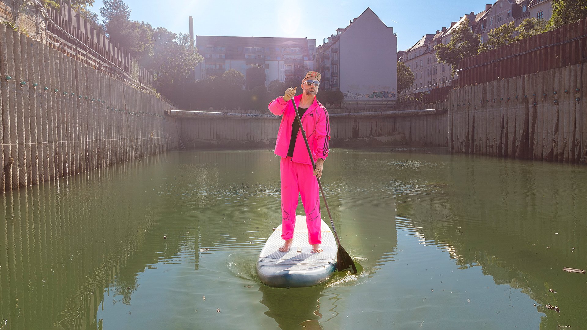 Foto für die Veranstaltung MC Harras – Alte, weiße Männer mit dem Künstler auf einem Paddelsurf-Board und im rosa Trainingsanzug.