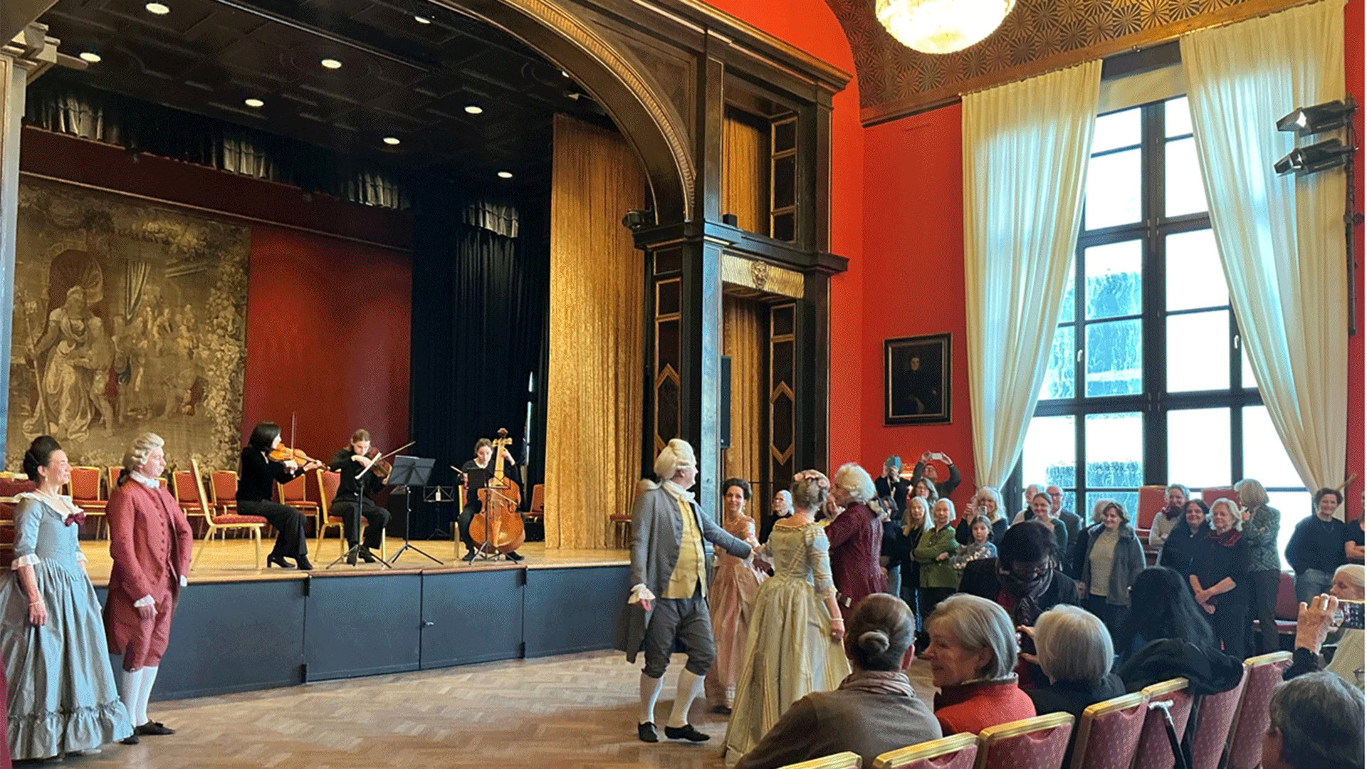 Historischer Tanzabend in einem festlich dekorierten Saal. Mehrere Personen in barocken Kostümen tanzen vor einer Bühne mit Streichensemble, während das Publikum auf Stuhlreihen und im Saal verteilt zuschaut.