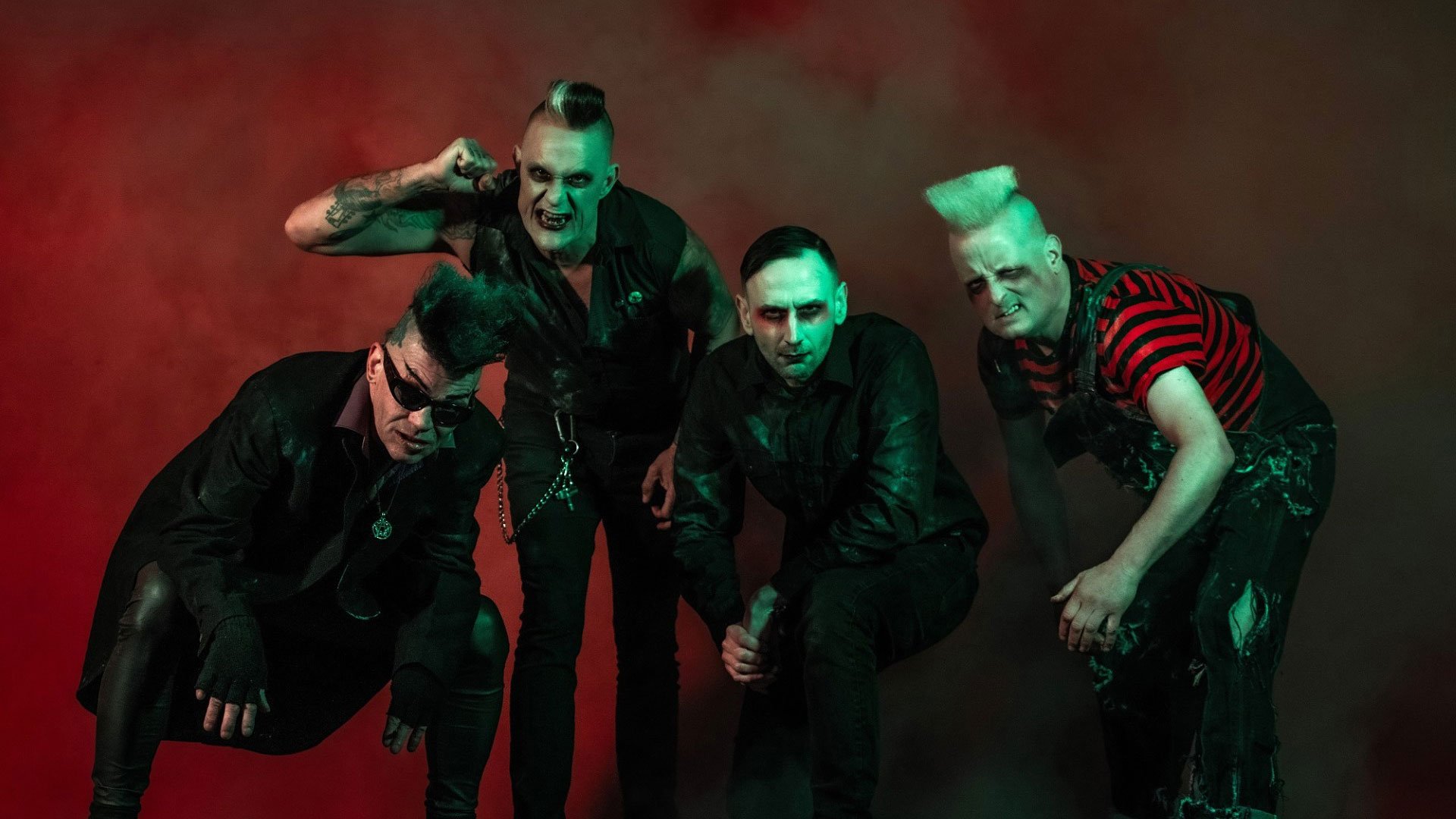 Vier Mitglieder der Band Demented Are Go posieren vor rot grün beleuchtetem Hintergrund. Schwarze Kleidung, auffällige Frisuren, Tätowierungen und kontrastreiche Bühnenbeleuchtung prägen die Gruppenaufnahme.