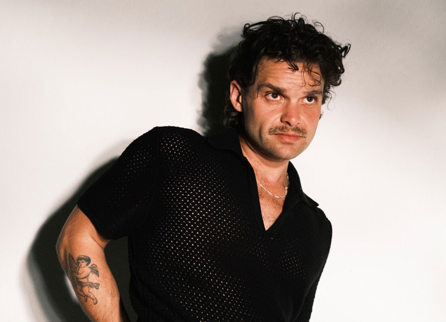 Ein Foto des Künstlers mit schwarzem Hemd, an eine weiße Wand gelehnt.