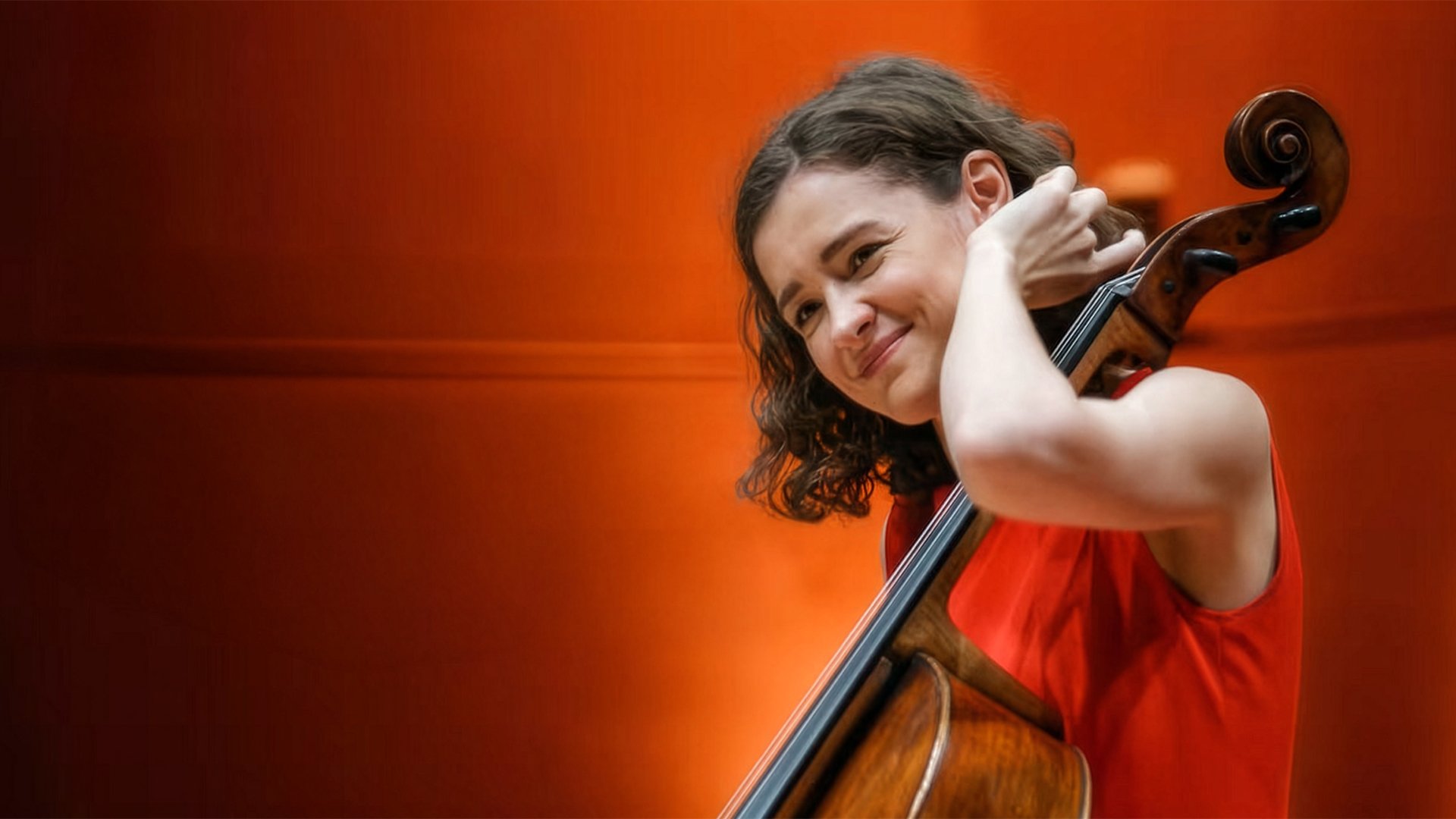 Musikerin mit Violoncello vor warm orangefarbenem Hintergrund, hält das Instrument nah am Körper.