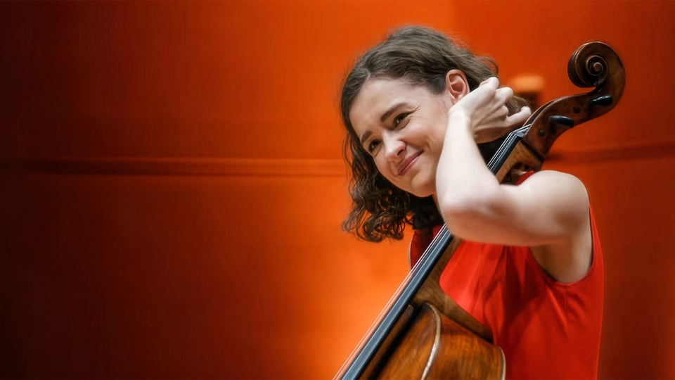 Musikerin mit Violoncello vor warm orangefarbenem Hintergrund, hält das Instrument nah am Körper.