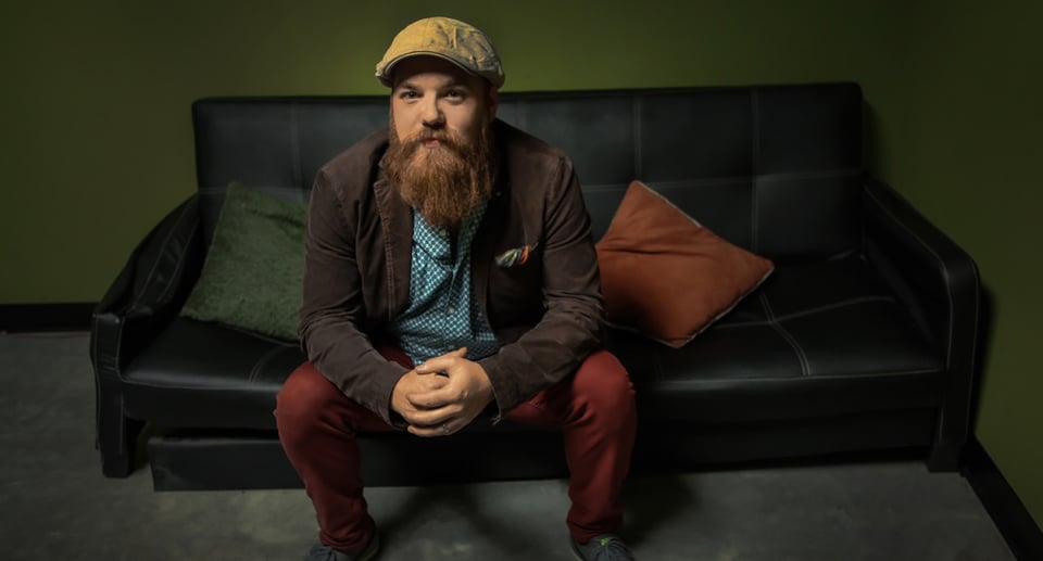 ein Mann mit langem Bart und Baskenmütze, der auf einem Sofa sitzt. Die Wand im Hintergrund ist grün