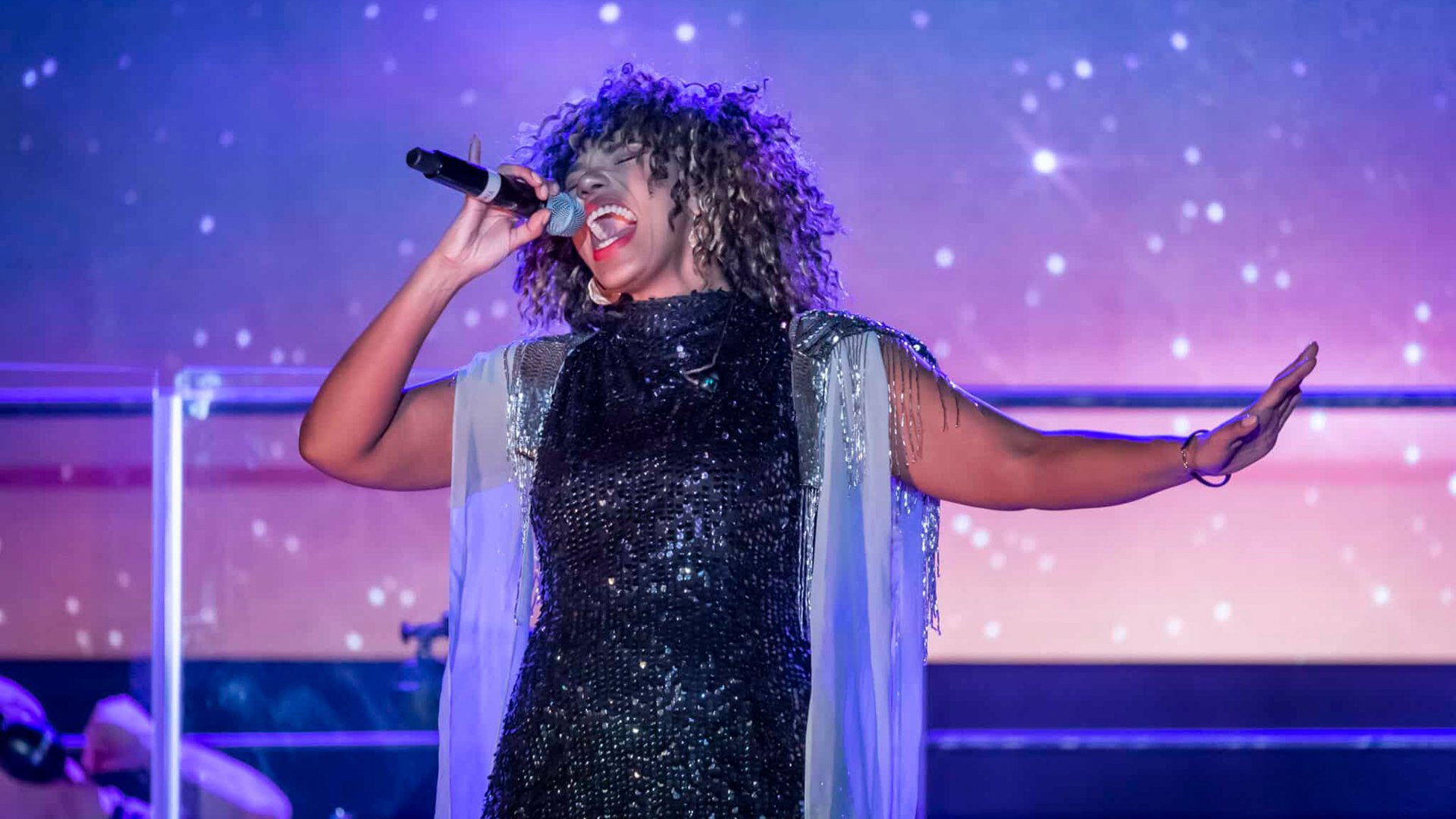 Darstellerin von TINA – The Rock Legend singt mit Mikrofon auf einer Konzertbühne vor farbig beleuchtetem Hintergrund.