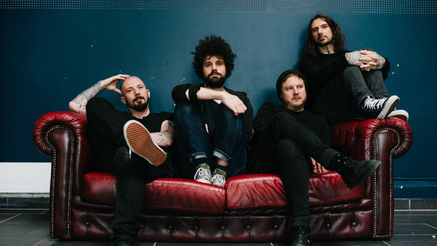 Die Band Coilguns sitzt auf einem antik wirkenden roten Ledersofa mit dunkelblauem Hintergrund