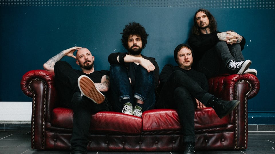 Die Band Coilguns sitzt auf einem antik wirkenden roten Ledersofa mit dunkelblauem Hintergrund