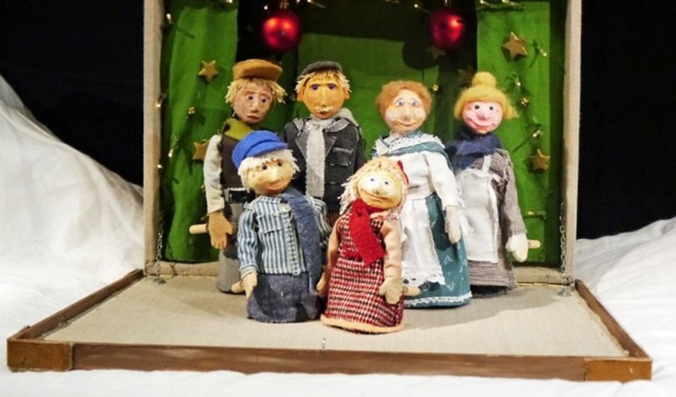 sechs Puppenfiguren stehend auf einem aufgeklappten Holzkoffer mit Weihnachtsdekoration im Hintergrund