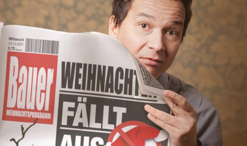 Person mit braunen Haaren und Blick in die Kamera hält eine Zeitung mit der Schlagzeile Weihnachten fällt aus Josef gesteht alles und einer durchgestrichenen Weihnachtsmütze vor sich, vor einem warm beleuchteten Hintergrund.
