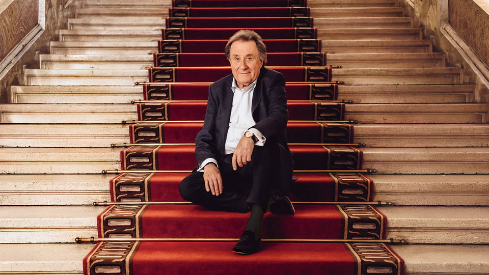 Rudolf Buchbinder in dunklem Anzug sitzt auf breiter, hellsteinener Treppe mit rotem Teppichläufer und goldenen Stangen in einem repräsentativen Innenraum