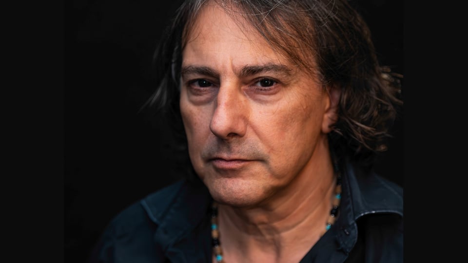 Ein Portrait des Künstlers Pippo Polina in Jeanshemd mit Kette vor dunklem Hintergrund