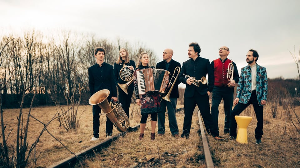 Sieben Musiker mit Instrumenten stehen auf einem Feld mit alten Gleisen