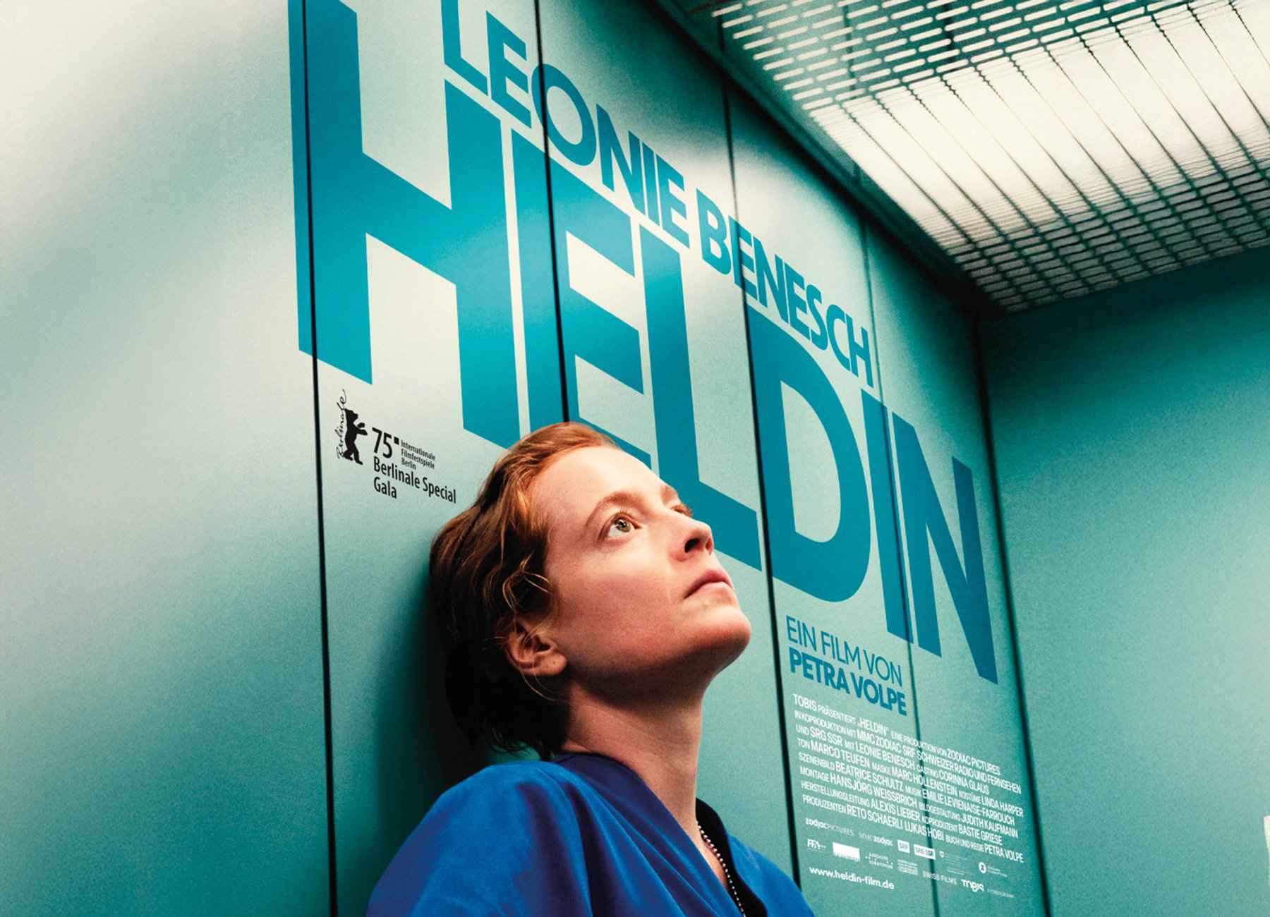 Dame in blauer Kleidung vor türkiser Wand mit Aufschrift des Filmtitels