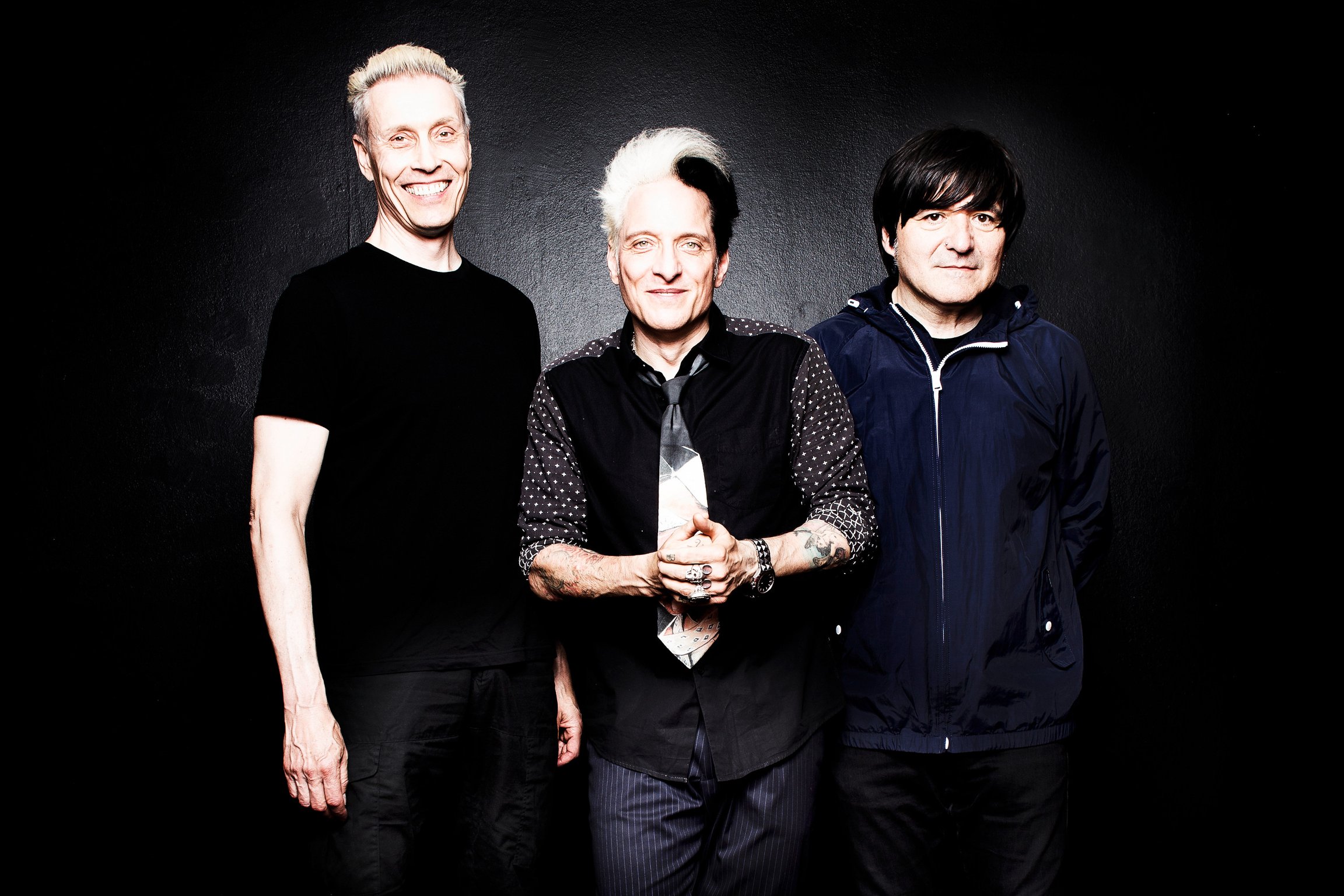 Die Ärzte gucken frontal in die Kamera und stehen vor einer dunklen Wand.