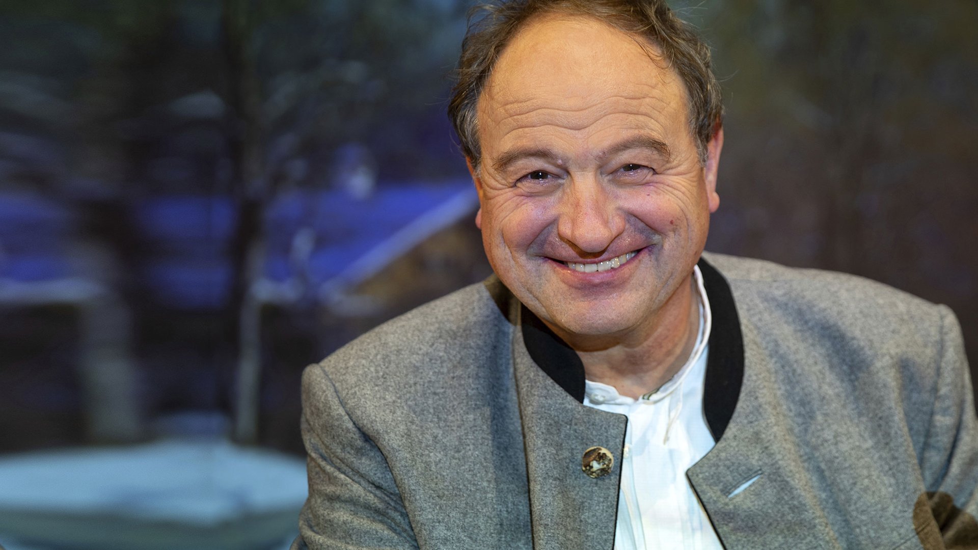 Porträt einer Person in traditioneller Trachtenjacke mit Knopfdetail, frontal aufgenommen vor unscharfem Hintergrund in dunklen und blauen Farbtönen