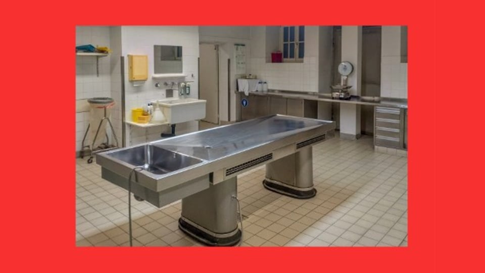 Edelstahl-Seziertisch in einem gekachelten Obduktionsraum mit Waschbecken, Arbeitsflächen und Waage, das Bild ist von einem breiten roten Rahmen umgeben, zur Veranstaltung Leichenpräparator Alfred Riepertinger Teil 2