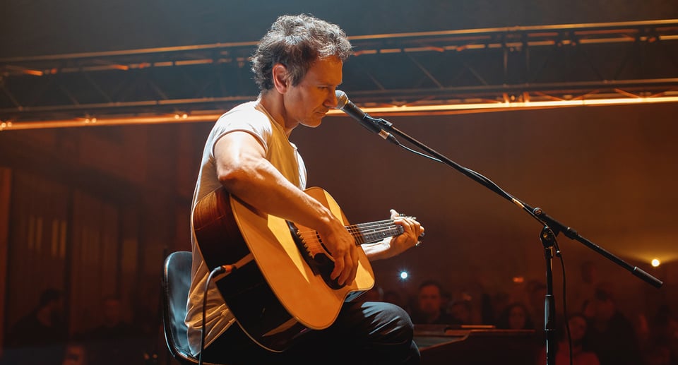 Foto des Künstlers „Slava Vakarchuk“ auf einer Bühne sitzend mit einer Gitarre und einem Mikrofon