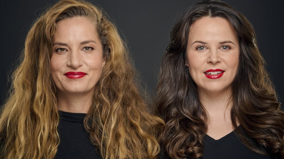 Zwei Damen mit langen Haaren und rotem Lippenstift in schwarzer Kleidung vor dunklem Hintergrund