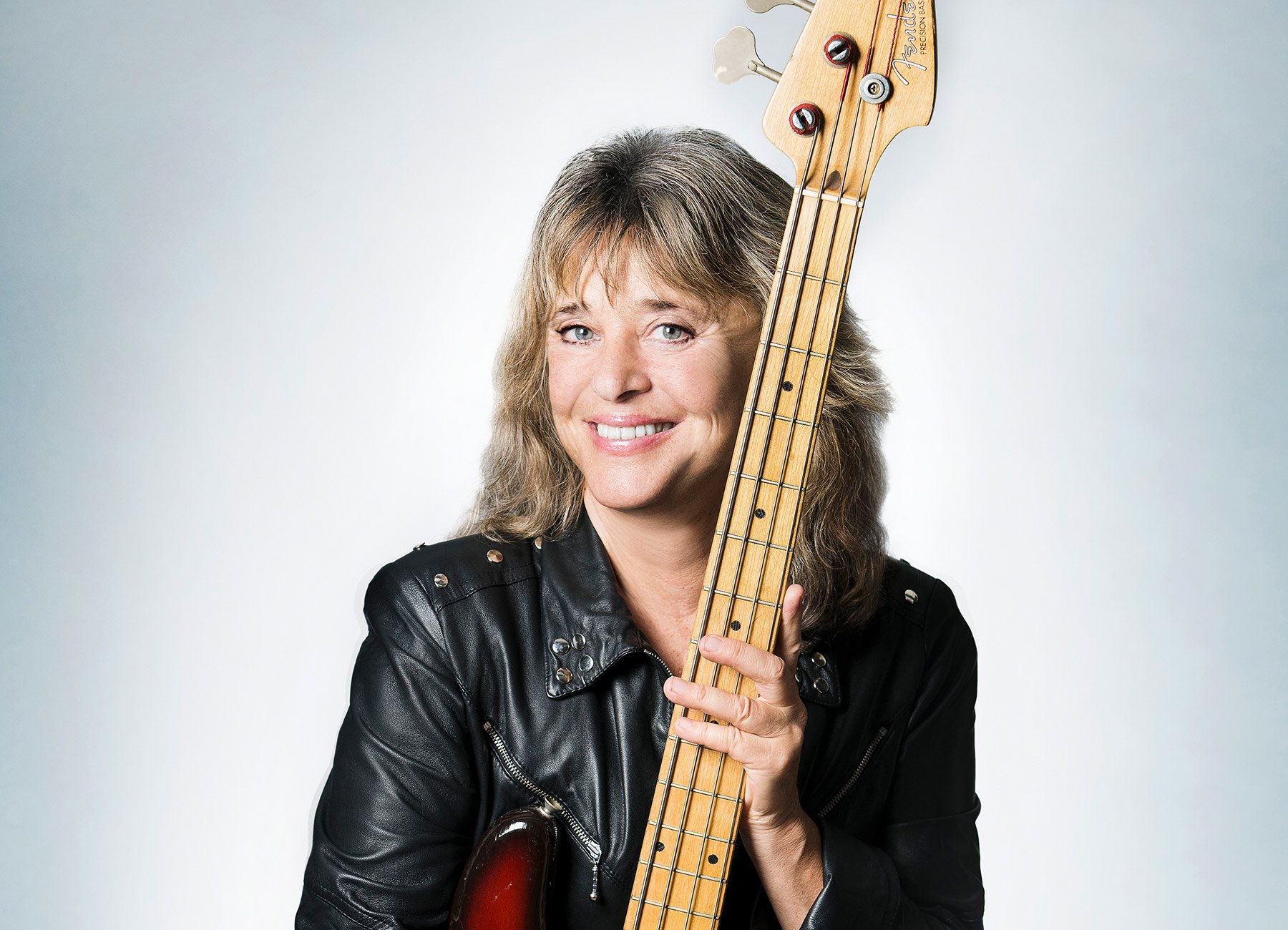 Foto der Künstlerin Suzi Quatro – WORLD-TOUR 2026, mit Lederjacke, Gitarre in der Hand, vor grauem Hintergrund