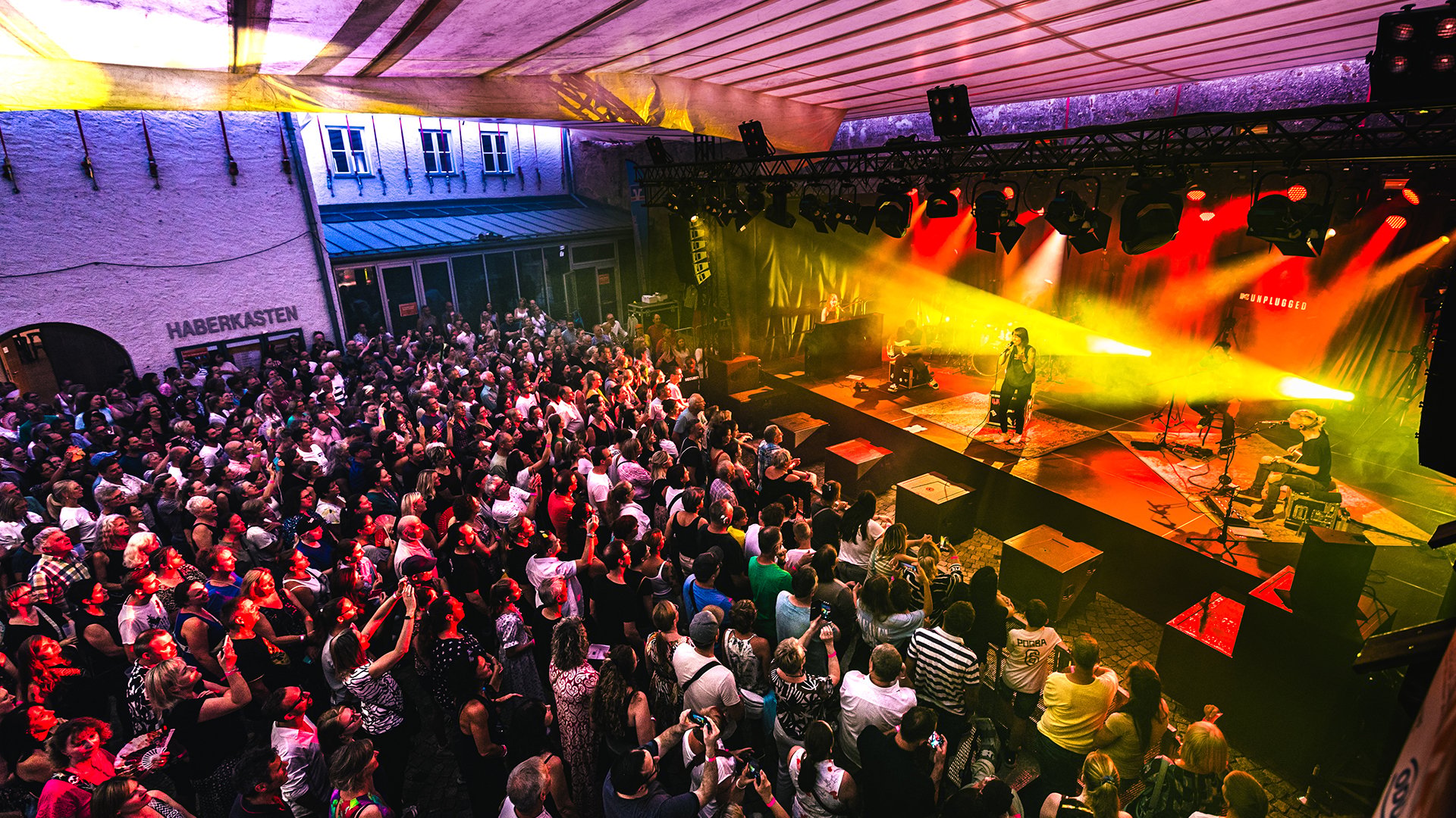 Große Menschenmenge bei Open-Air-Konzert vor Bühne mit Lichtshow, dichtes Publikum und Festivalatmosphäre in urbanem Innenhof