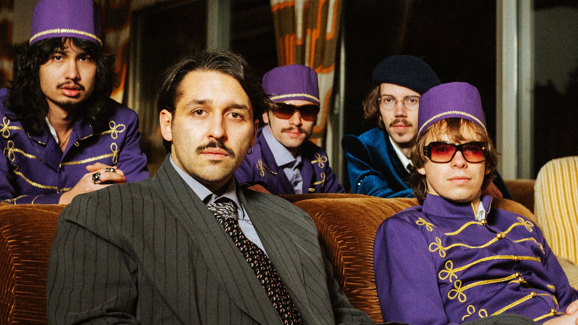 Foto für die Veranstaltung ‚TIAVO – Hotel Tiavo Tour 2026‘ – die Gruppe sitzt auf Sofa und Sesseln, drei von ihnen in lila Anzügen und mit Hüten