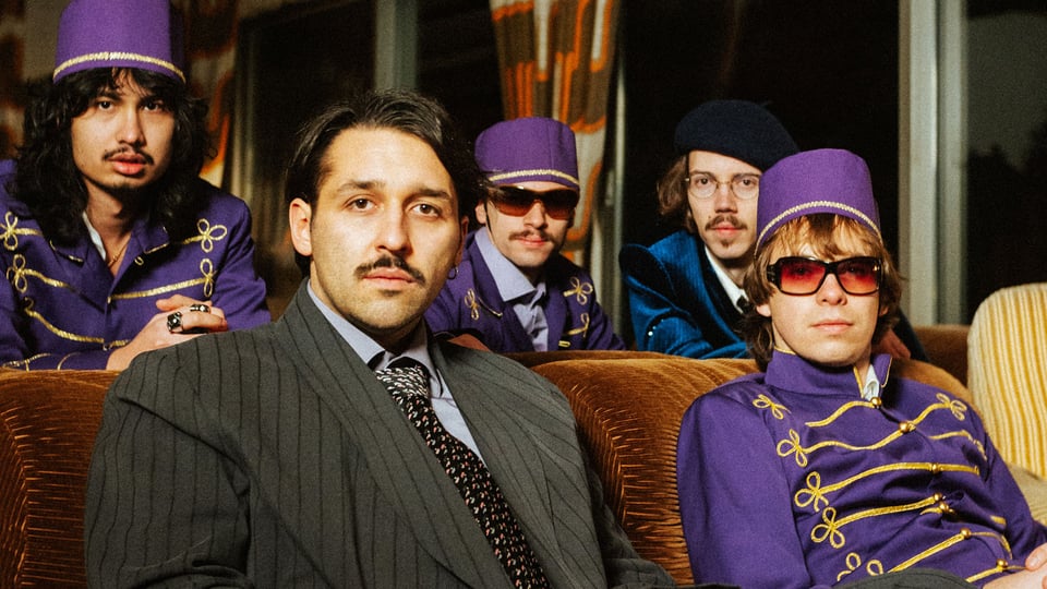 Foto für die Veranstaltung ‚TIAVO – Hotel Tiavo Tour 2026‘ – die Gruppe sitzt auf Sofa und Sesseln, drei von ihnen in lila Anzügen und mit Hüten