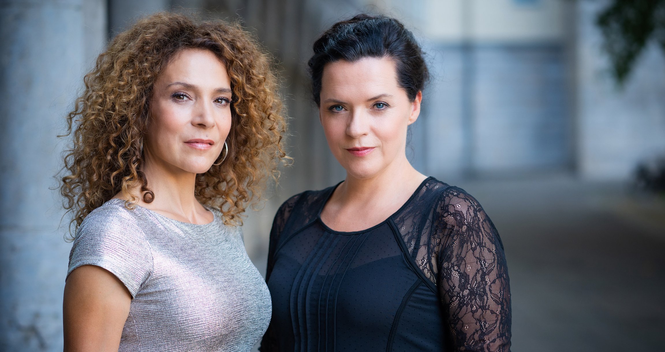 Zwei Damen, eine mit mittelblonden langen Locken eine mit dunklen hochgesteckten Haaren