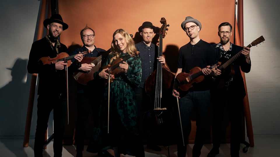 Die sechsköpfige Band mit Instrumenten vor einer orangefarbenen Leinwand