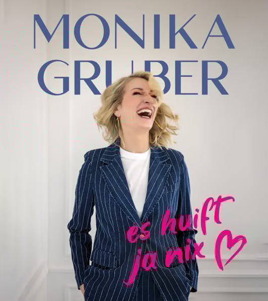 Monika Gruber lacht in einem dunkelblauen, weiß-gestreiftem Jeansanzug mit der Aufschrift "es huift ja nix" in pink.