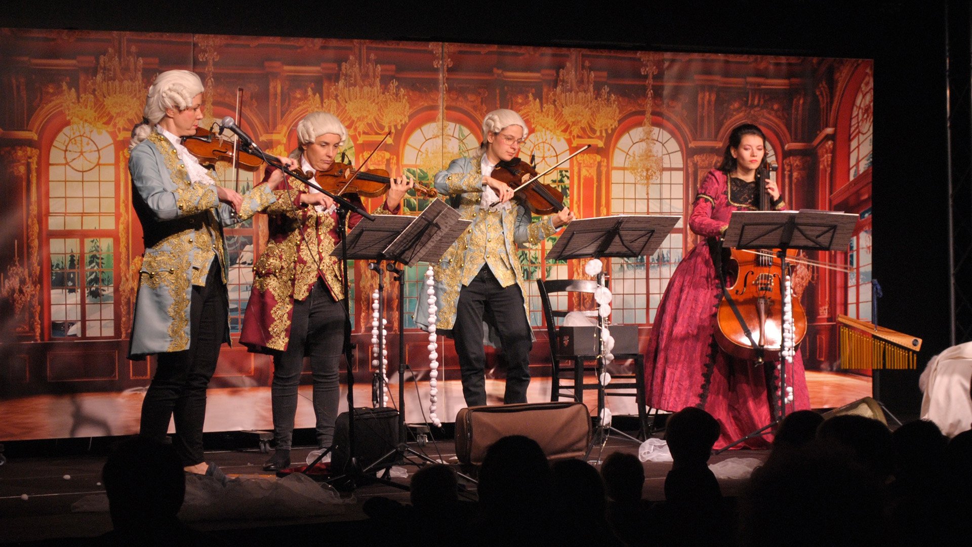 Vier Musikerinnen und Musiker in historischen Kostümen des 18. Jahrhunderts spielen auf einer Bühne Violinen und Violoncello. Die Bühne ist mit einer Ballsaal-Kulisse im barocken Stil gestaltet, im Vordergrund sind Notenständer und die Silhouetten des Publikums zu sehen.