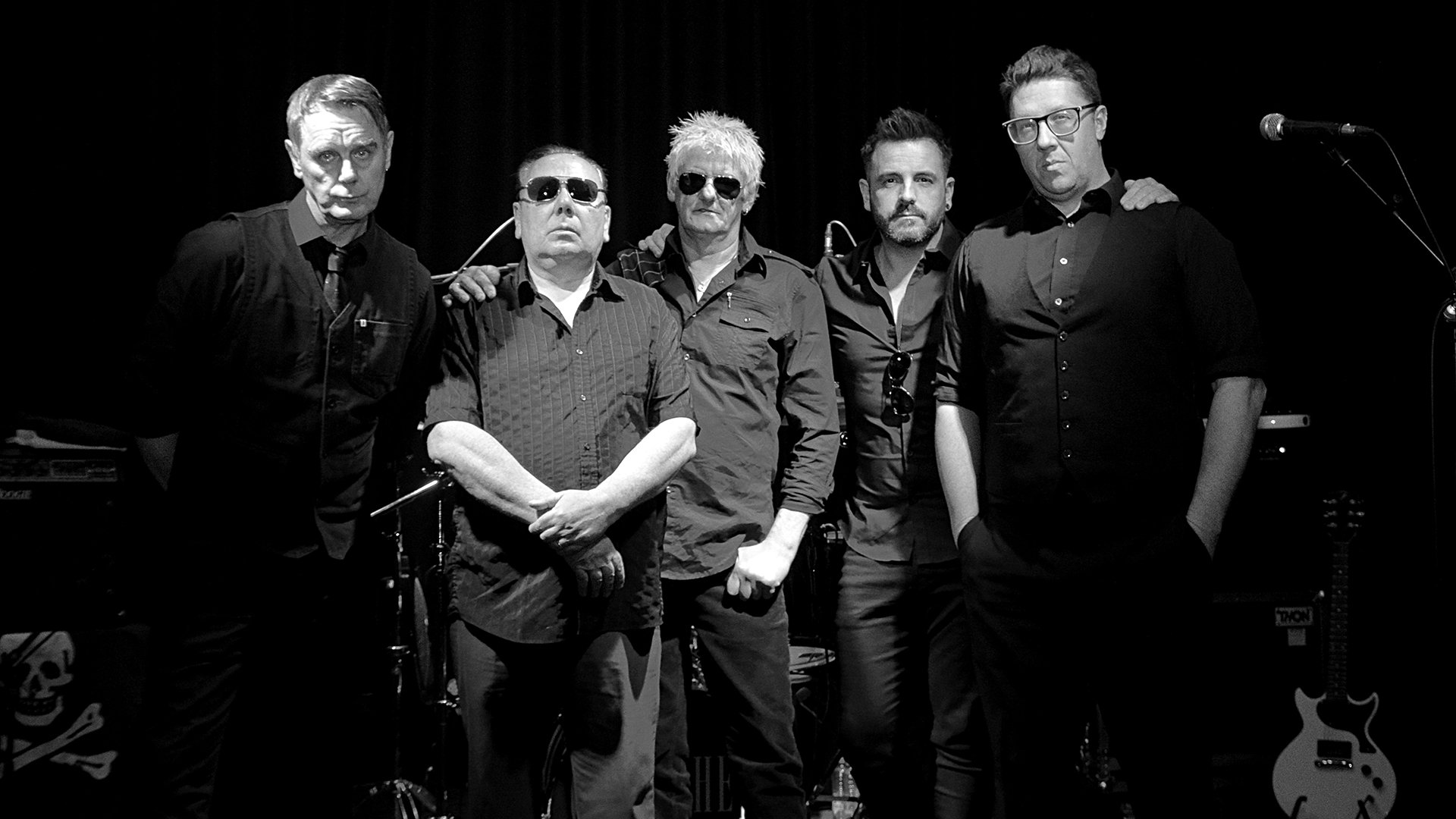 Bandfoto von The Godfathers mit fünf Bandmitgliedern in dunkler Kleidung auf einer Bühne vor schwarzem Hintergrund, zwei Personen mit Sonnenbrillen im Zentrum, umgeben von Mikrofonständern, Schlagzeug und E-Gitarre in Schwarzweißaufnahme.