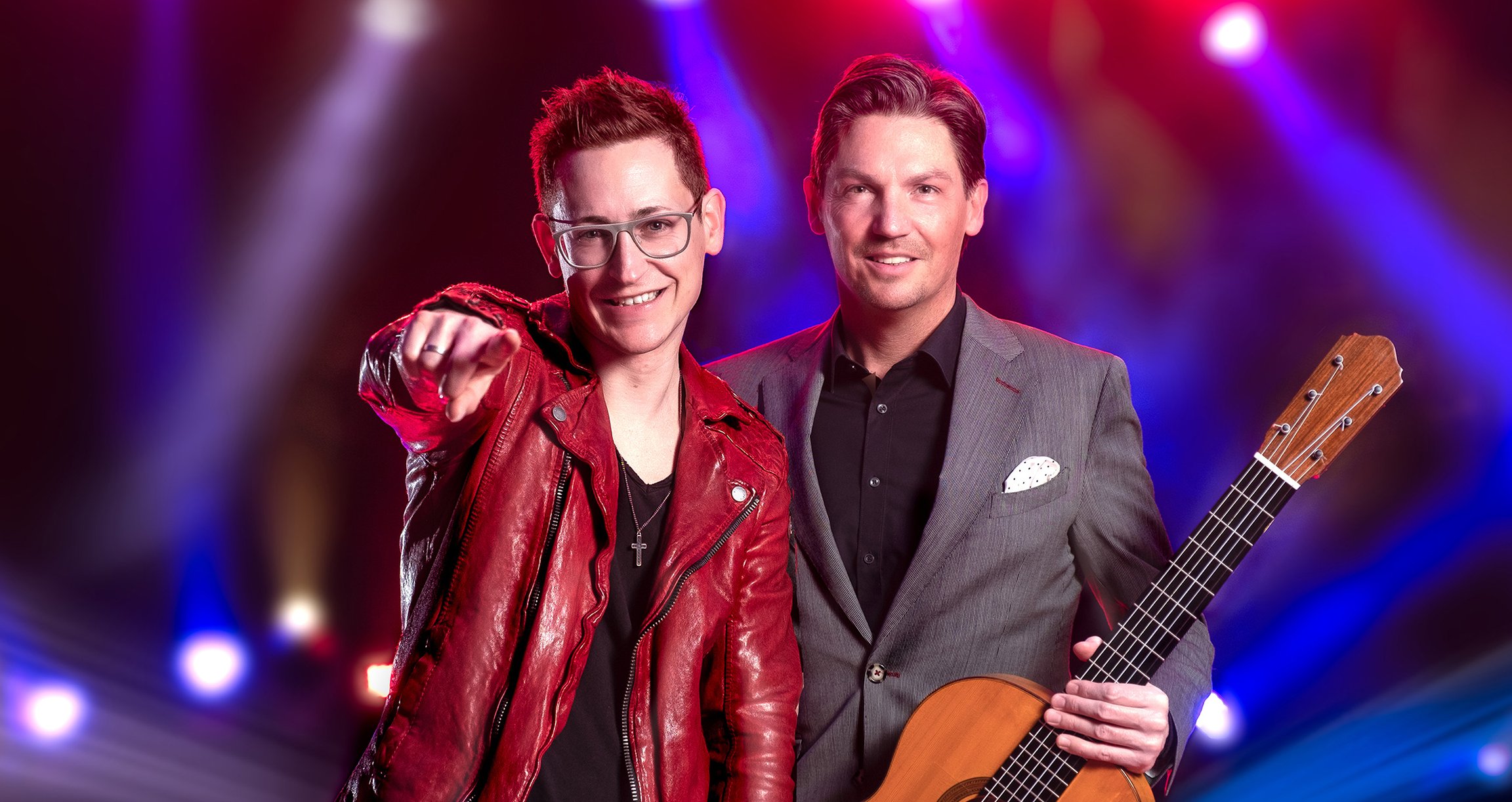 Zwei Männer, einer mit Gitarre in grauem Anzug, einer mit Brille und roter Lederjacke vor buntem Scheinwerferlicht