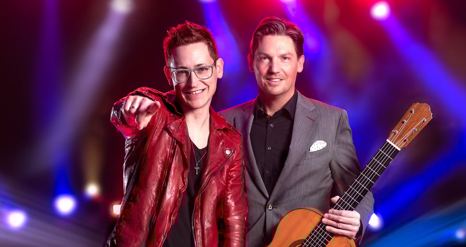Zwei Männer, einer mit Gitarre in grauem Anzug, einer mit Brille und roter Lederjacke vor buntem Scheinwerferlicht